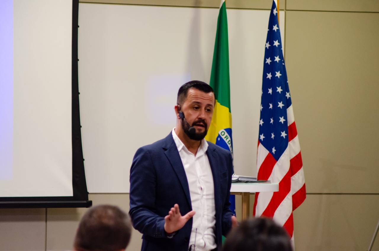 Cristovão Lucena palestrando com bandeiras do Brasil e EUA ao fundo